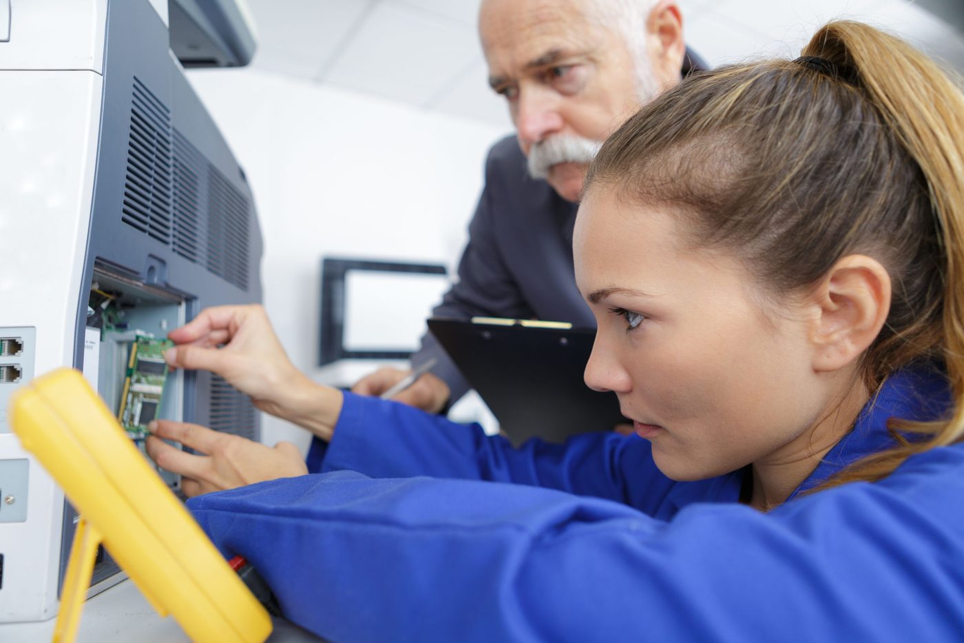 A senior coach and a junior technician in blue coveralls jointly diagnose a circuit board from an industrial machine — the transmission of knowledge through analysis at the heart of the DIAGFIAB© action-learning program